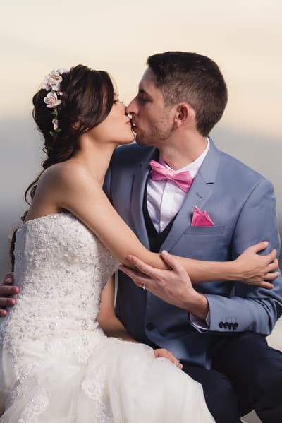 The bride Wing and groom Jason share a kiss during their couple portraits session. Wing wears a strapless white lace wedding gown with floral hair accessories, and Jason wears a light blue suit with a pink bow tie and pocket square. Engagement party by She Said Yes.