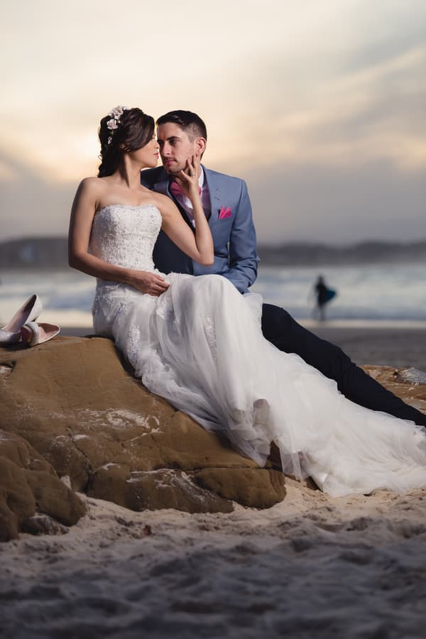 The bride Wing in a strapless white wedding gown and the groom Jason in a blue suit with a pink pocket square sit closely together on a rock at Bilinga Beach Weddings during a couple portrait session at sunset.