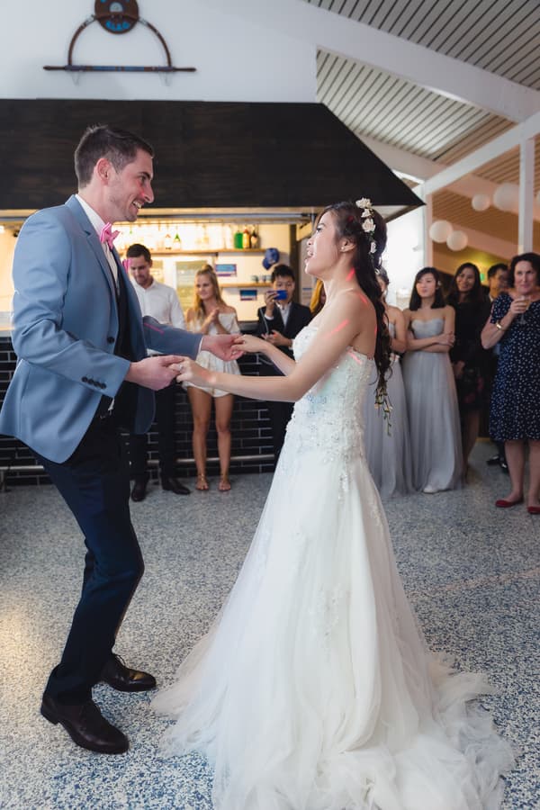 Bride Wing and groom Jason hold hands and dance on the reception stage at Bilinga Beach Weddings — Bilinga SLSC, while guests and bridesmaids watch in the background.