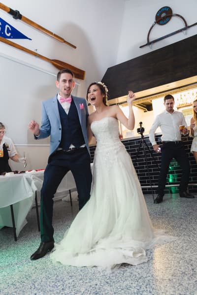Bride Wing and groom Jason dance together on the reception stage, with guests and a server visible in the background near a table and bar area. Winter wedding light photography Brisbane by She Said Yes.