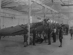 A line of airplanes is being assembled under the watchful eye of a gun-toting soldier as they are being completed to be sent to the European war; the factory is at College Point