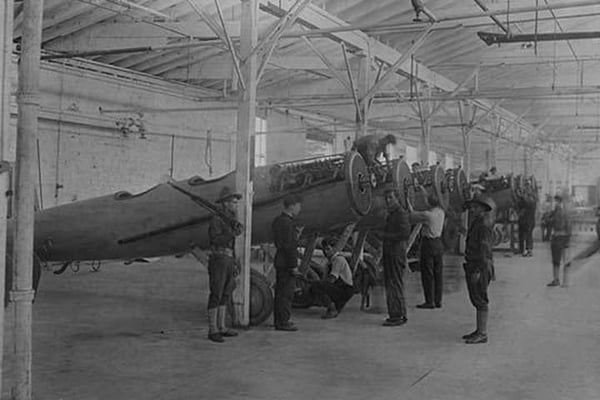A line of airplanes is being assembled under the watchful eye of a gun-toting soldier as they are being completed to be sent to the European war; the factory is at College Point