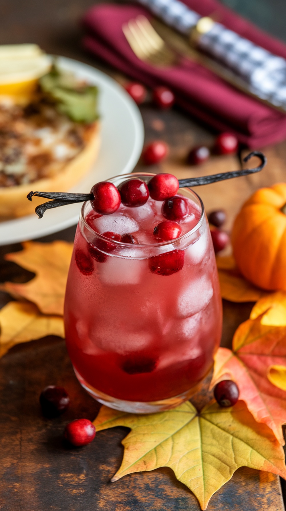 A cranberry vanilla mocktail garnished with cranberries and a vanilla pod, surrounded by autumn leaves and a pumpkin.