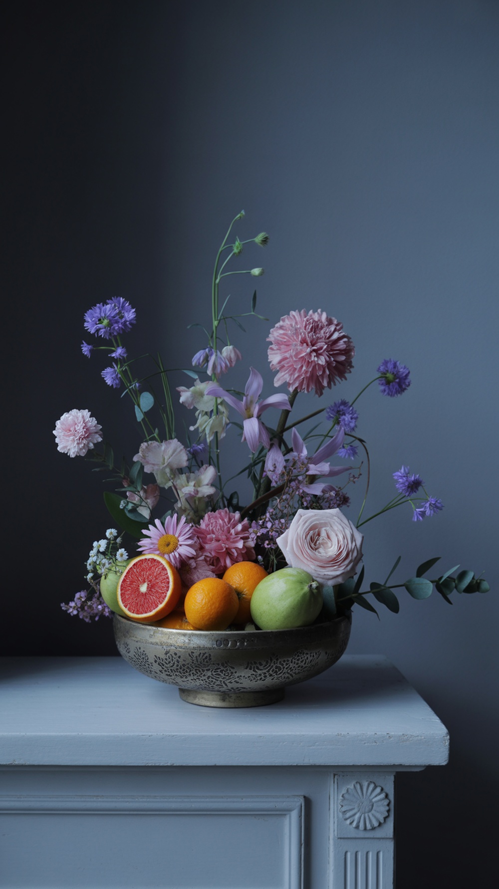 A beautiful arrangement of flowers and fruits in a decorative bowl, showcasing pink roses, purple flowers, oranges, and grapefruit.