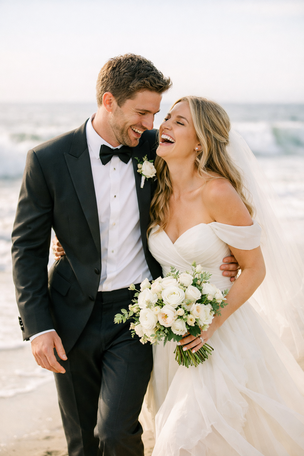 A happy couple laughing together on the beach during an engagement shoot, with the bride holding a bouquet and the groom in a tuxedo.