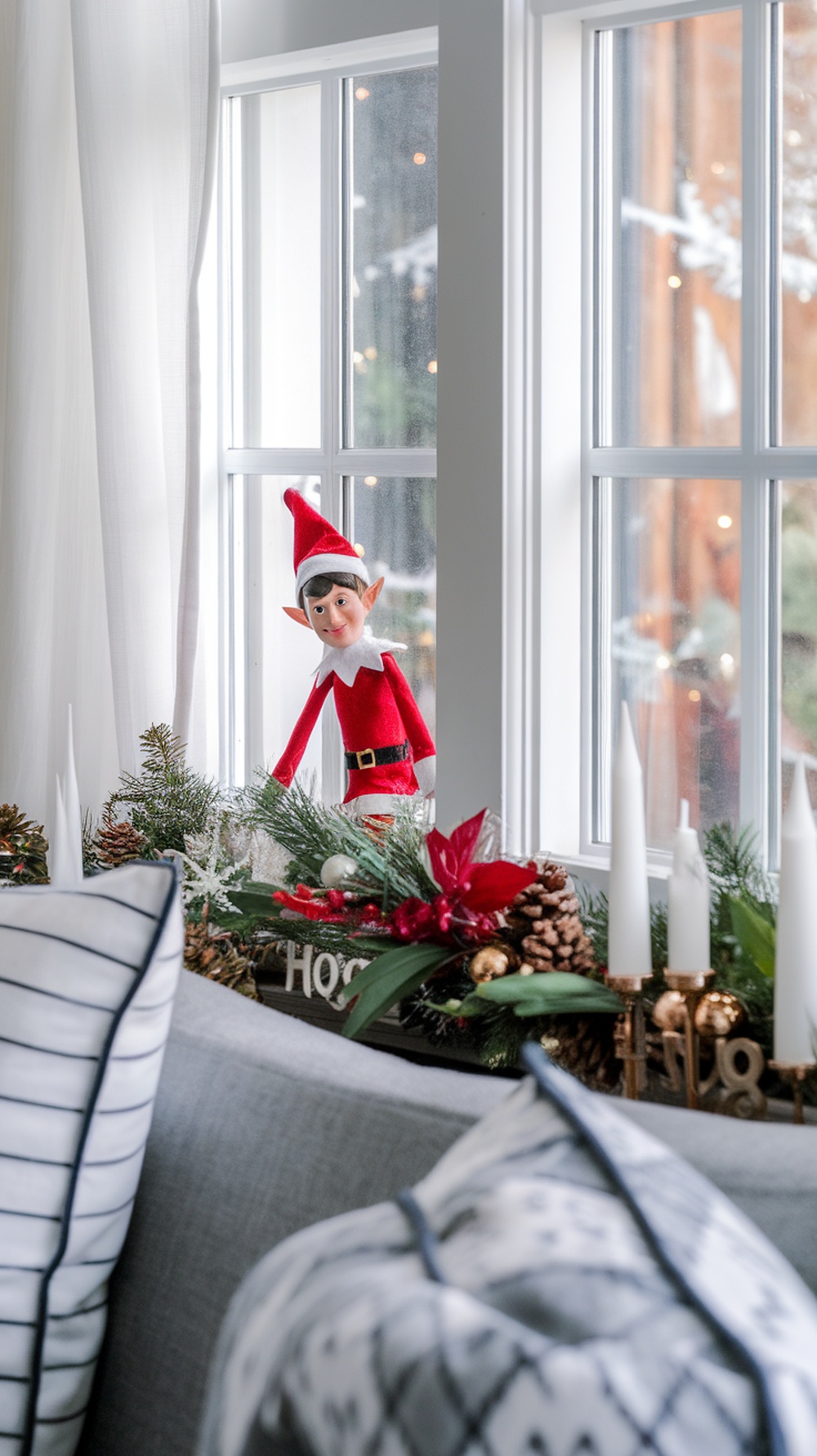 A whimsical elf figurine in a red outfit with a pointy hat, peeking through a window decorated with greenery and festive items.