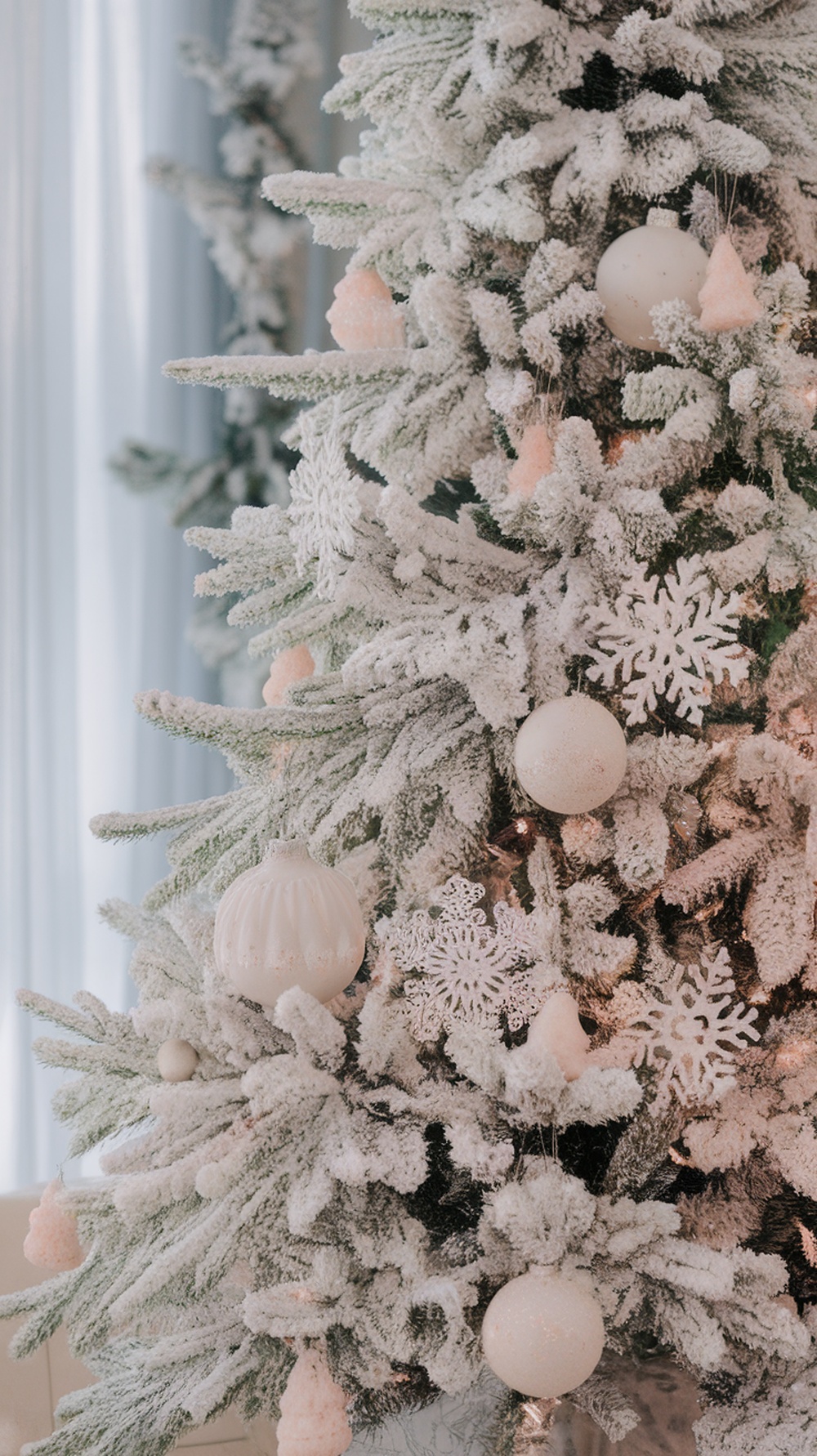 A frosted white Christmas tree decorated with white and pastel ornaments, including snowflakes and small pink trees.