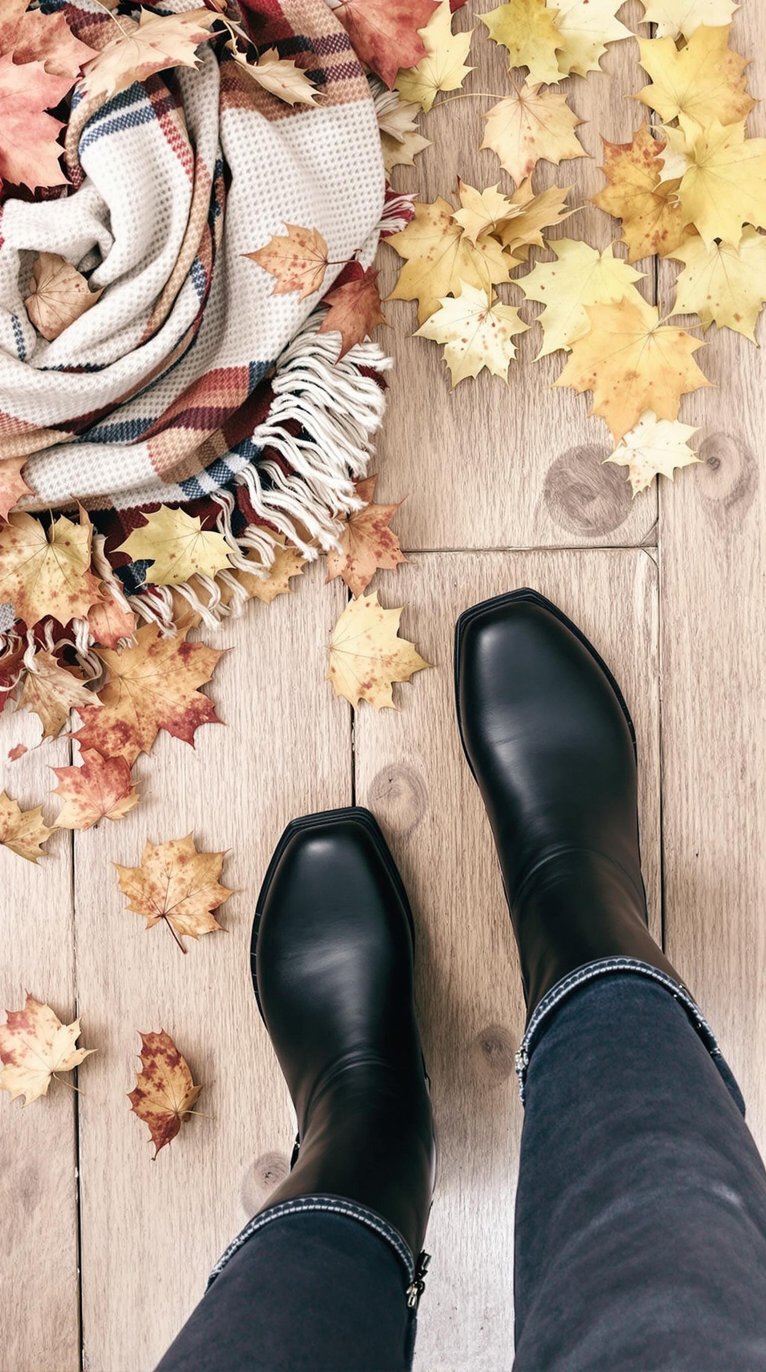 A pair of black Chelsea boots on a wooden surface surrounded by autumn leaves and cozy scarves.
