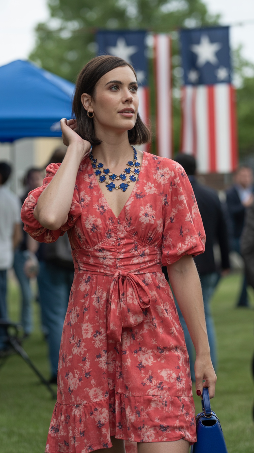 A woman in a red floral dress with blue accessories, posing outdoors with American flags in the background.