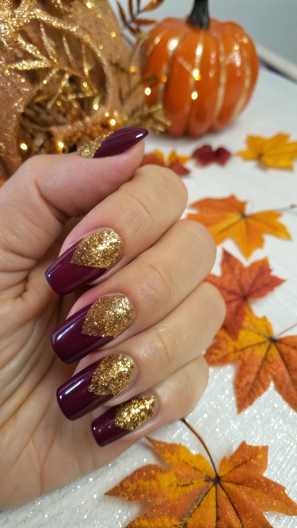 Nails painted in plum with gold glitter accents, surrounded by autumn leaves and a pumpkin.