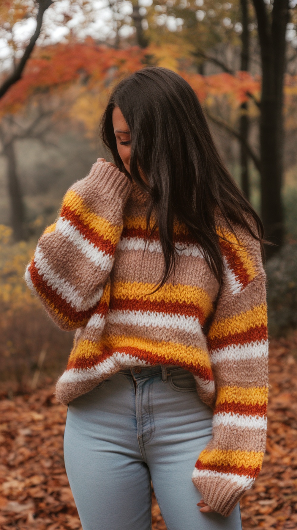 A person wearing a chunky knit sweater with a tree design and high-waisted jeans, surrounded by autumn leaves.