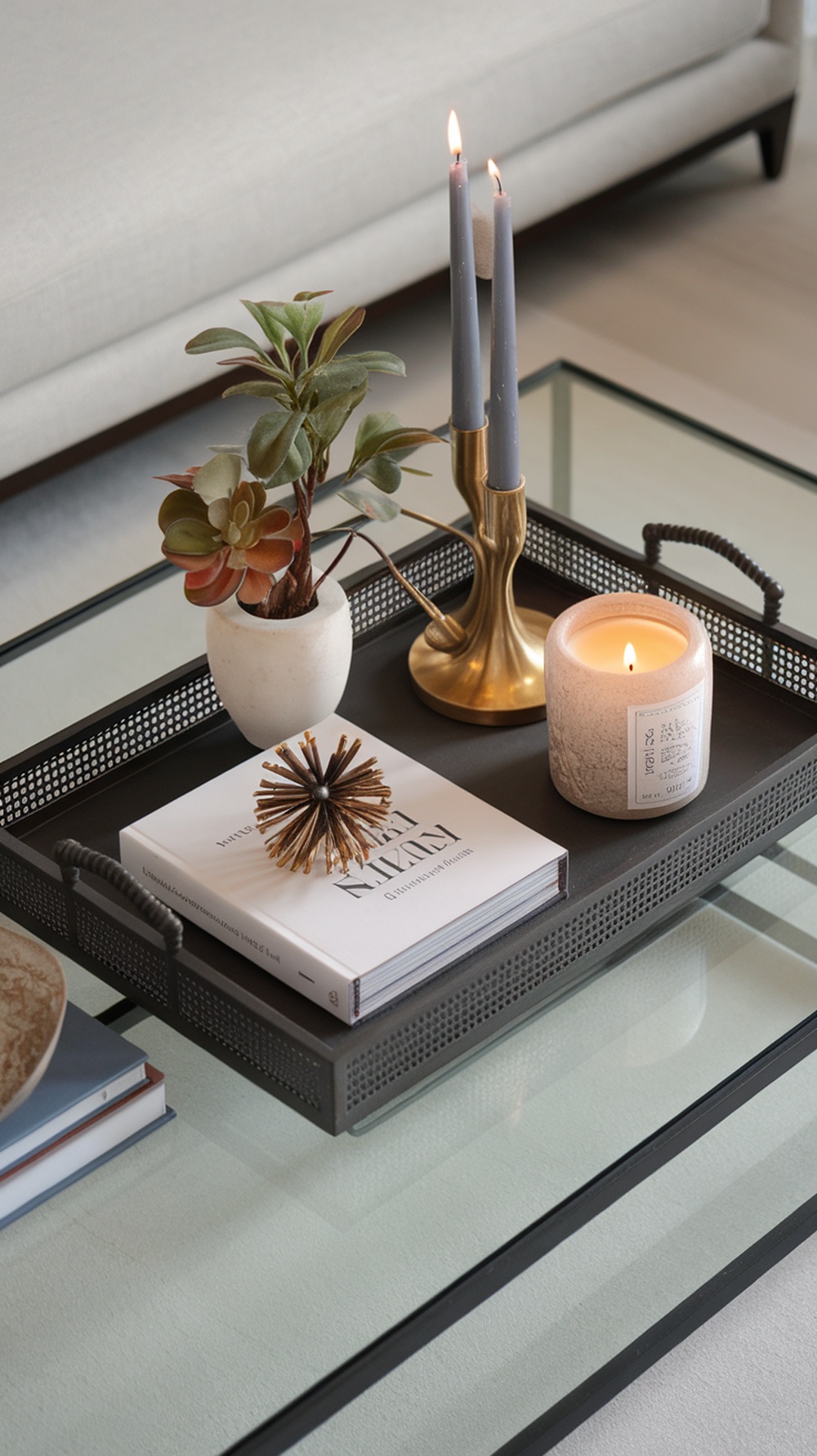 A decorative black tray with candles, a plant, a decorative object, and a book on a glass table.