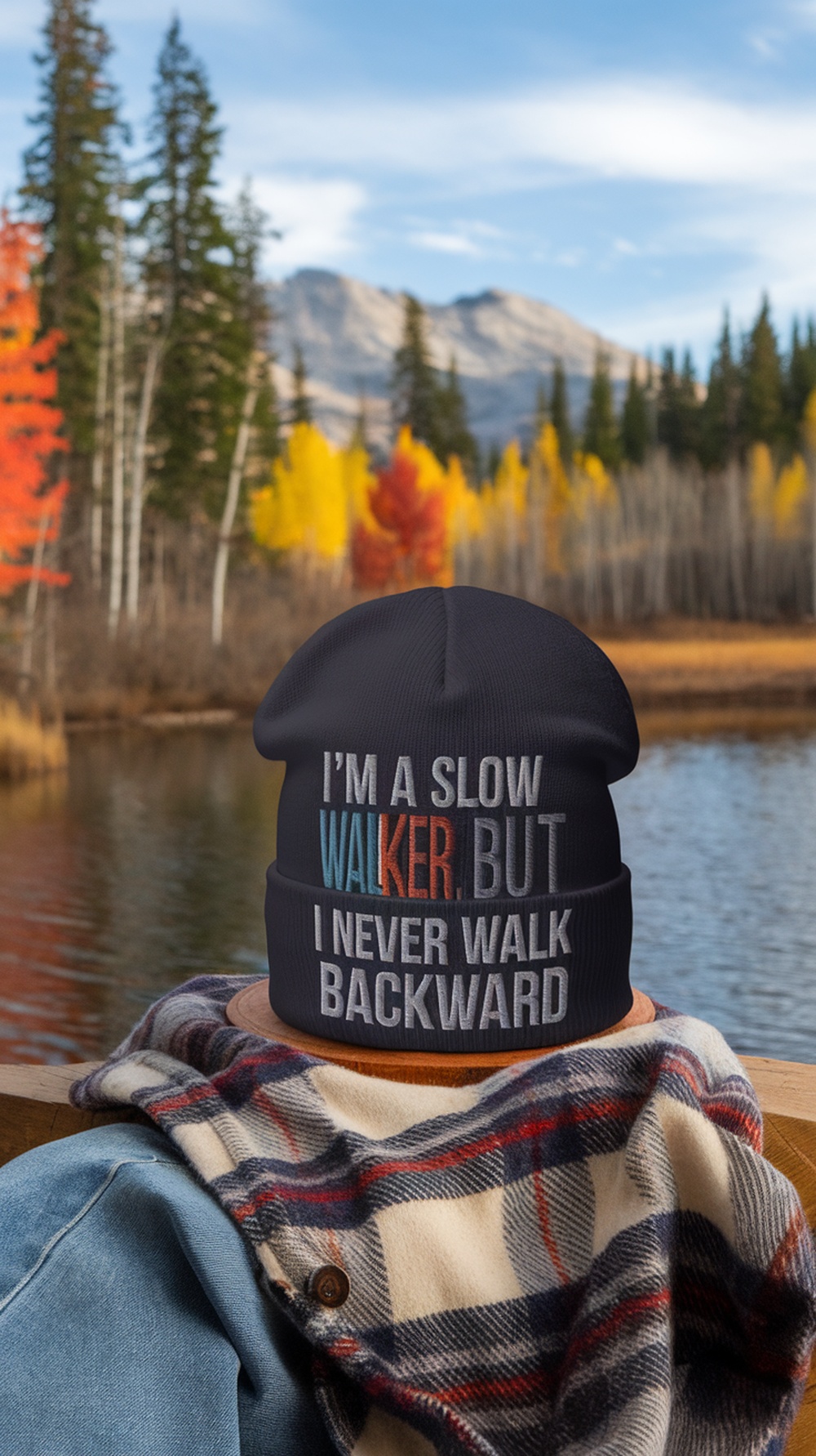 A person wearing a knit beanie with a humorous phrase, sitting by a lake surrounded by autumn foliage.