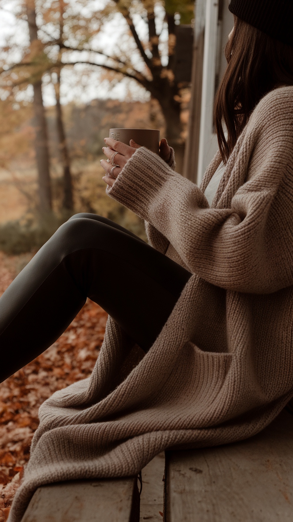 A person sitting on a porch wearing an oversized cardigan and leggings, holding a warm drink, with autumn leaves in the background.