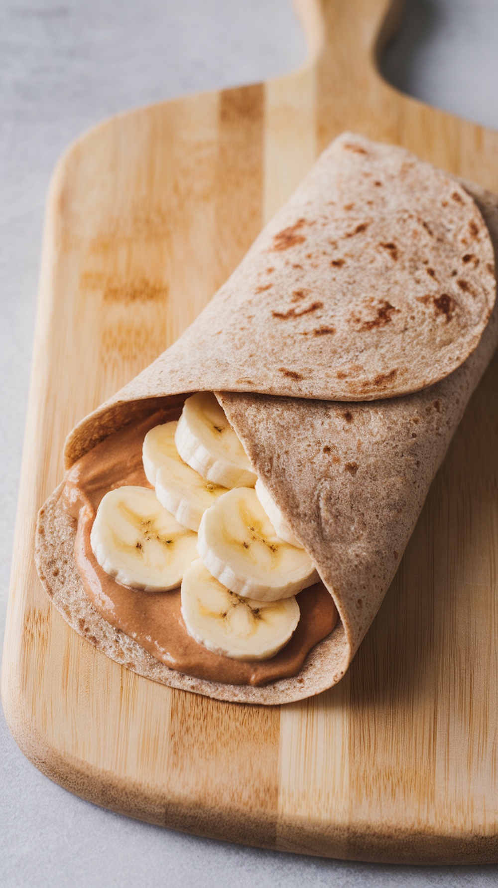 A whole grain wrap filled with nut butter and banana slices, resting on a wooden board.