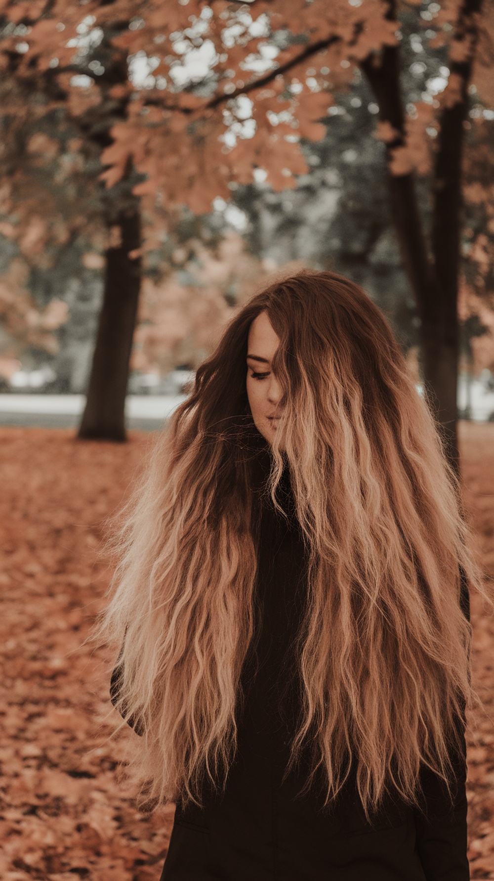 A woman with long, curly hair styled in layers with an ombre effect, standing in a fall setting with orange leaves.