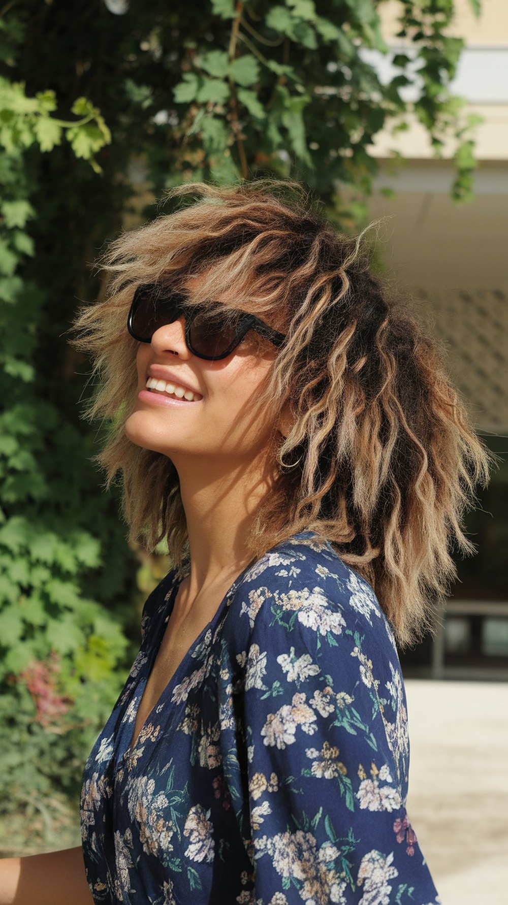 A woman with curly hair styled in textured layers, wearing sunglasses and a floral outfit, smiling outdoors.