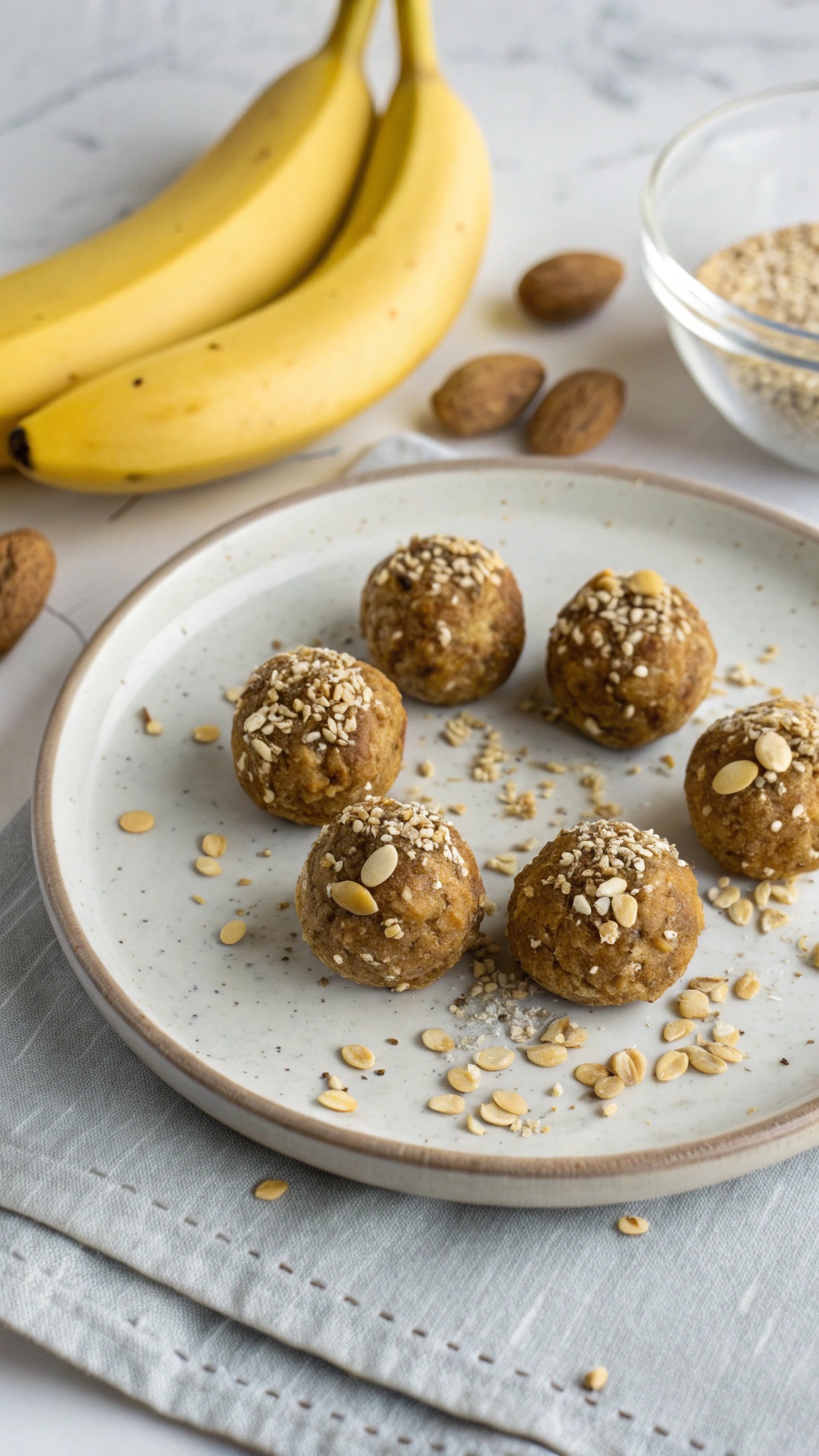 A plate of banana peanut butter energy balls sprinkled with oats, surrounded by fresh bananas and almonds.