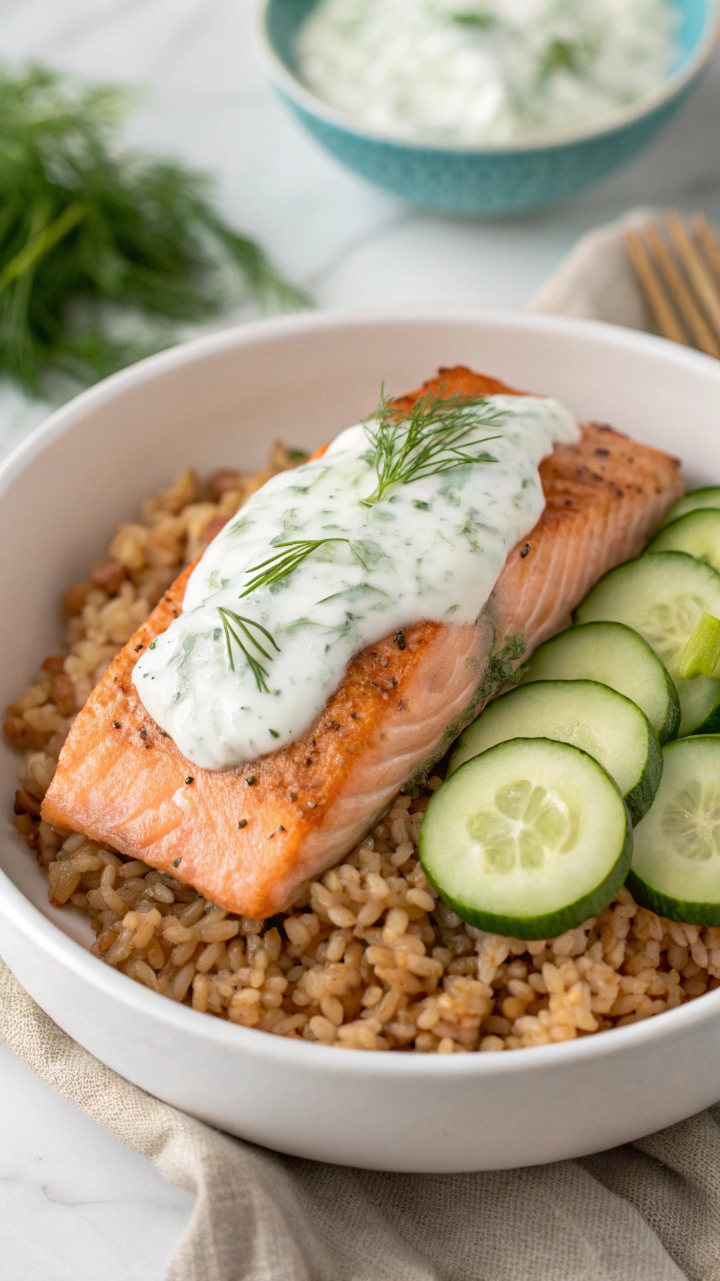 A Greek Yogurt Dill Salmon Bowl with salmon, dill sauce, brown rice, and cucumber slices.