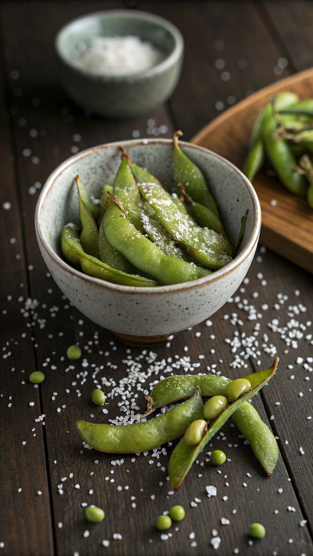 A bowl of steamed edamame sprinkled with sea salt, with some edamame pods and salt scattered on a wooden surface.