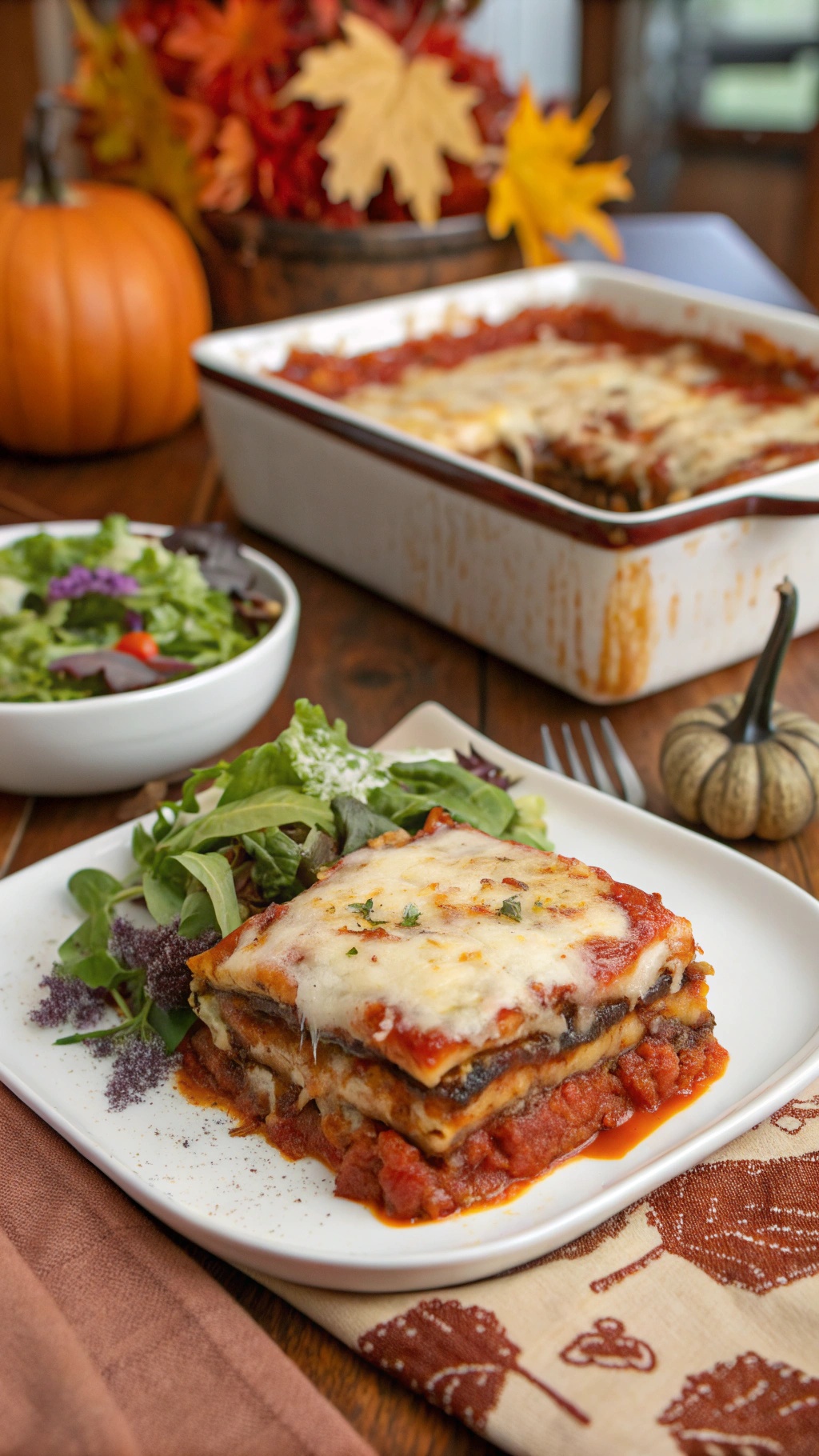 A plate of eggplant lasagna with a side salad, set on a wooden table with fall decorations.