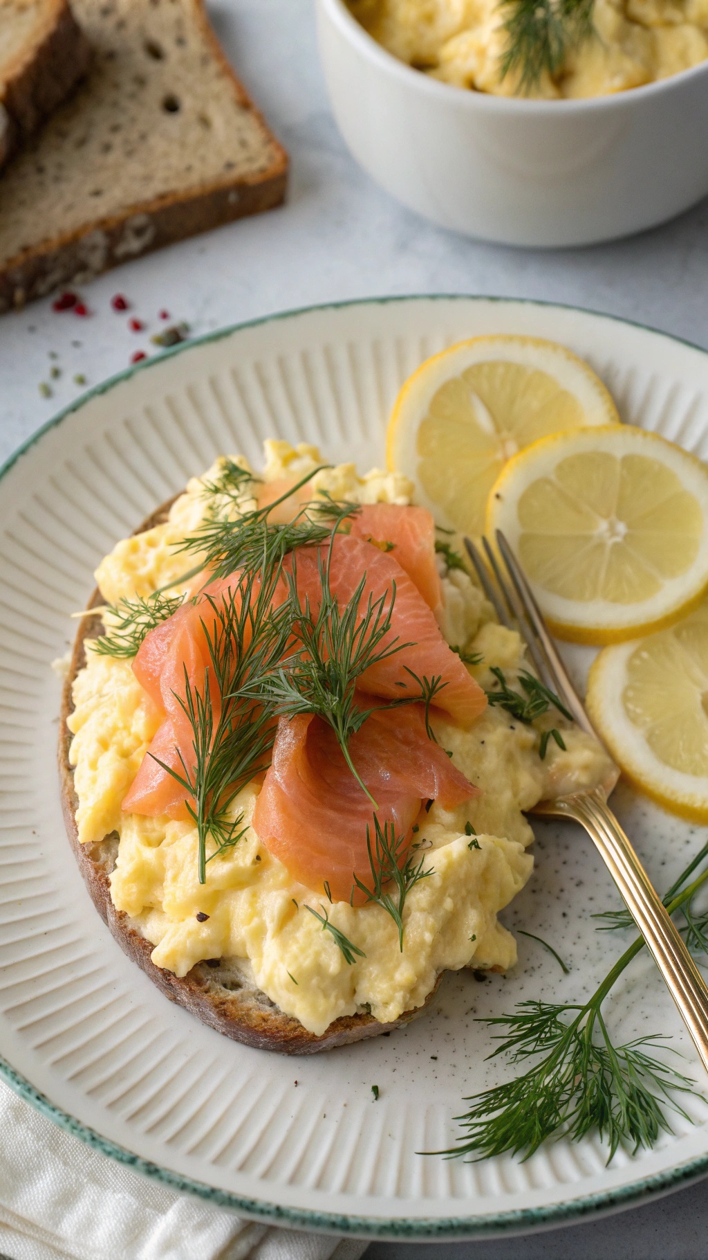 A plate of scrambled eggs topped with smoked salmon, fresh dill, and lemon slices, served on whole-grain bread.