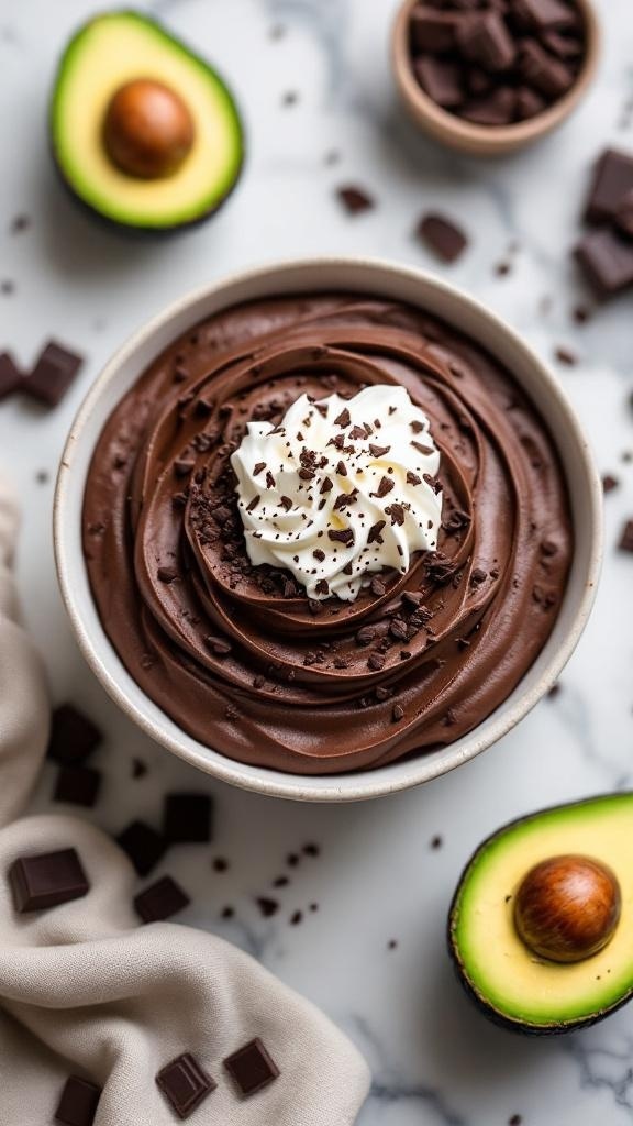 A bowl of dark chocolate avocado mousse topped with whipped cream and chocolate shavings, surrounded by avocado halves and chocolate pieces.