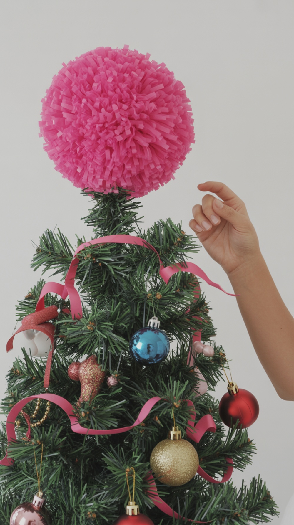 A hand placing a pink pom-pom topper on a Christmas tree decorated with colorful ornaments.
