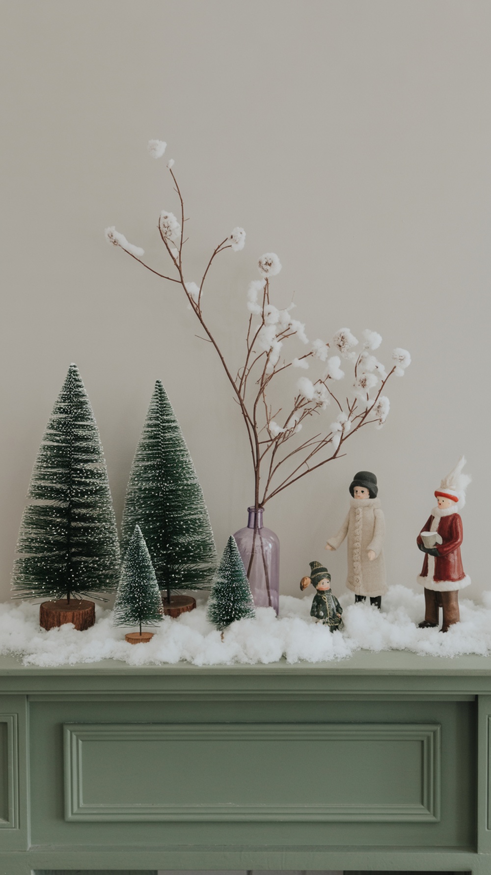 A decorated fireplace mantel featuring small evergreen trees, winter figurines, and fake snow.