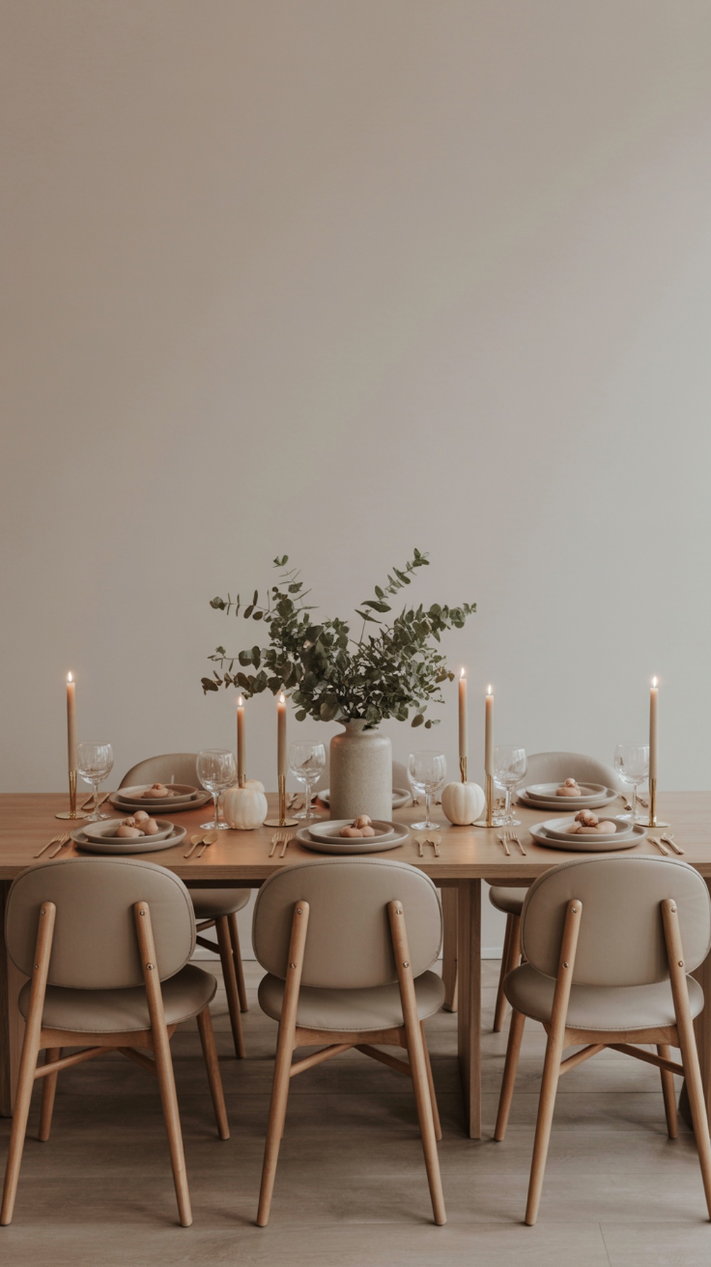 A minimalist Thanksgiving table setting with a wooden table, neutral plates, glassware, eucalyptus centerpiece, and white pumpkins.