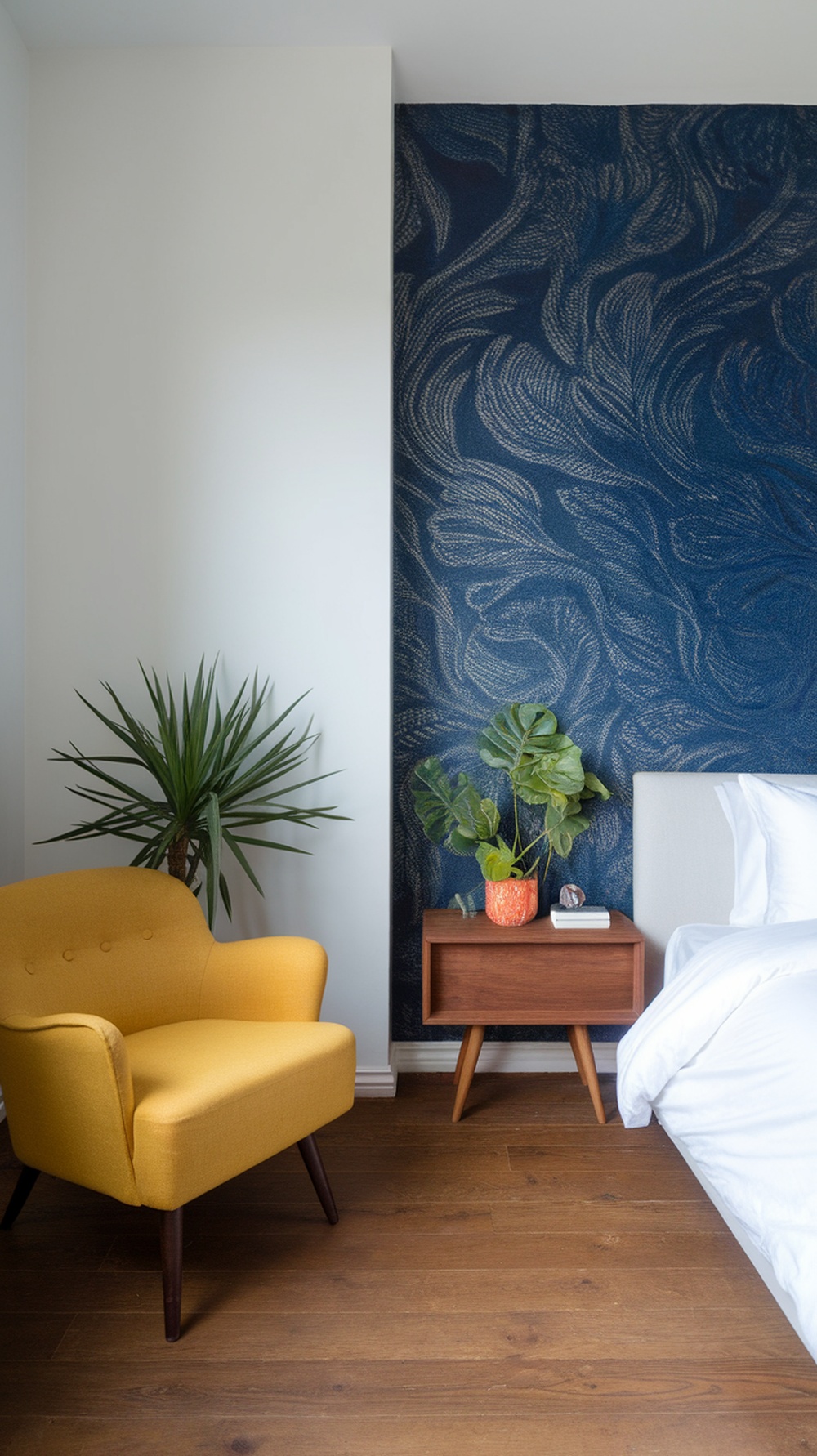 A stylish bedroom featuring a deep blue patterned wall, a bright yellow chair, a wooden nightstand, and a potted plant.