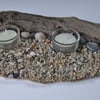 Twin glass tealight holder & tealight on pebble beach with a driftwood backdrop.