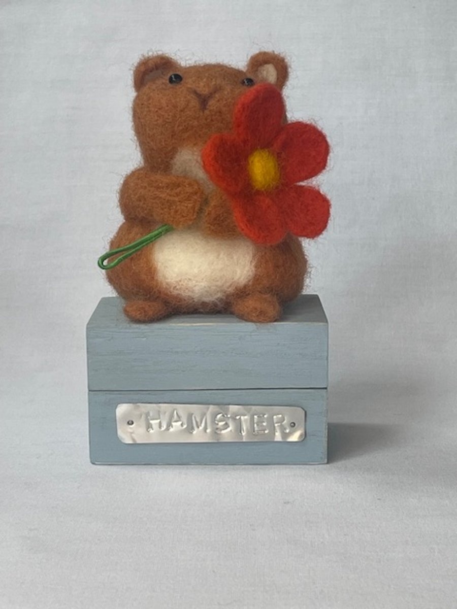 Handcrafted needle felted hamster holding a flower on a wooden box