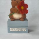 Handcrafted needle felted hamster holding a flower on a wooden box