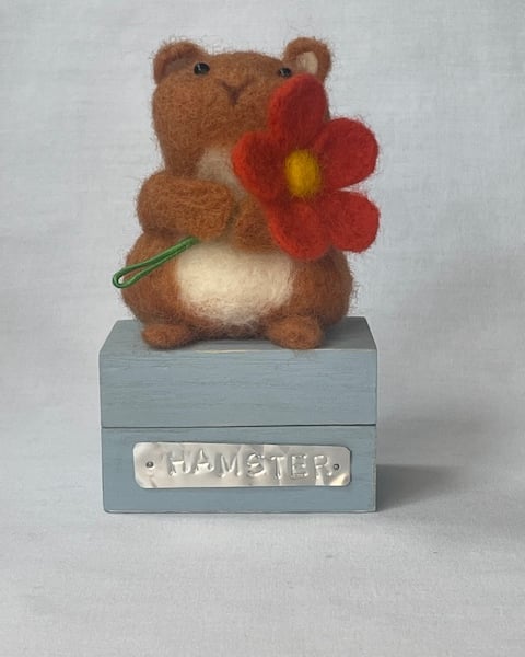 Handcrafted needle felted hamster holding a flower on a wooden box
