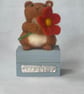 Handcrafted needle felted hamster holding a flower on a wooden box