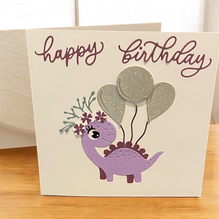 Smiling Dinosaur Birthday Card – with a floral headbands and balloons 