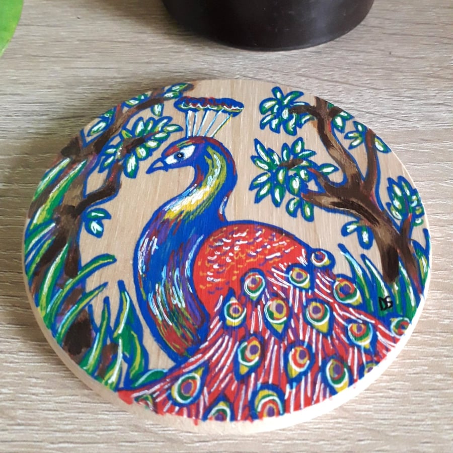 Circular wooden coaster hand drawn with a red and blue peacock among the trees. 