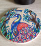 Circular wooden coaster hand drawn with a red and blue peacock among the trees. 