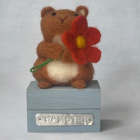 Handcrafted needle felted hamster holding a flower on a wooden box