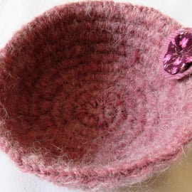 A small crochet basket or bowl in fluffy pink yarn with a fabric rosette 