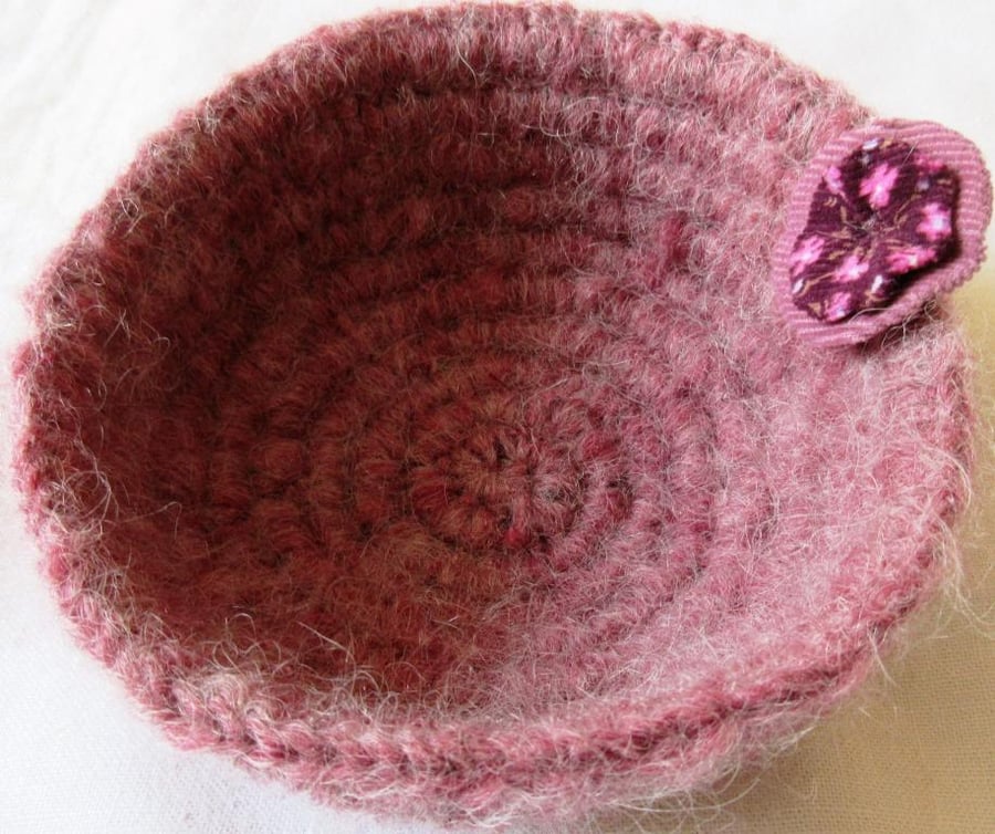 A small crochet basket or bowl in fluffy pink yarn with a fabric rosette 