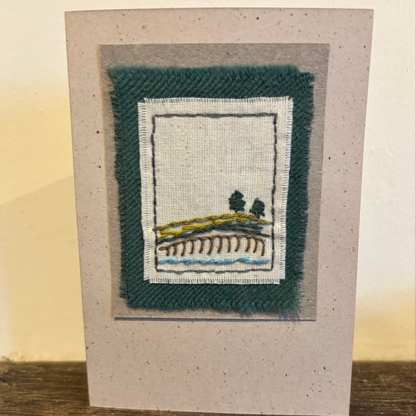 Handmade card featuring embroidered landscape of pine trees by river bank