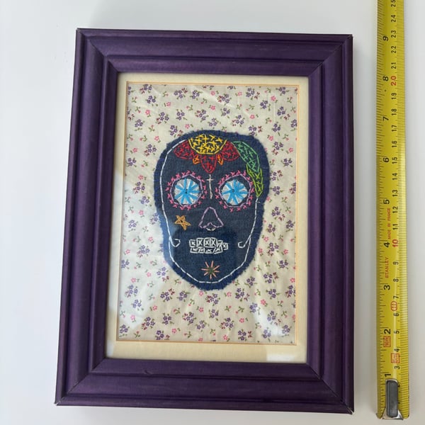 Textile art sugar skull in a purple frame.