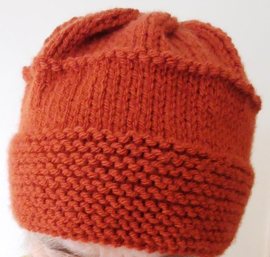 A hand-knitted beanie-style woman's hat in chunky acrylic yarn in a rust colour 