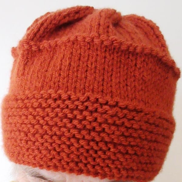 A hand-knitted beanie-style woman's hat in chunky acrylic yarn in a rust colour 