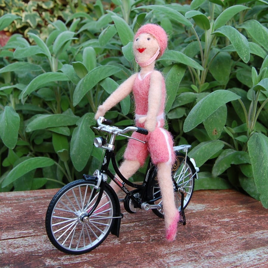 Going for a ride.  Needlefelt figure dressed in pink sitting on a metal cycle 