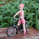 Going for a ride.  Needlefelt figure dressed in pink sitting on a metal cycle 