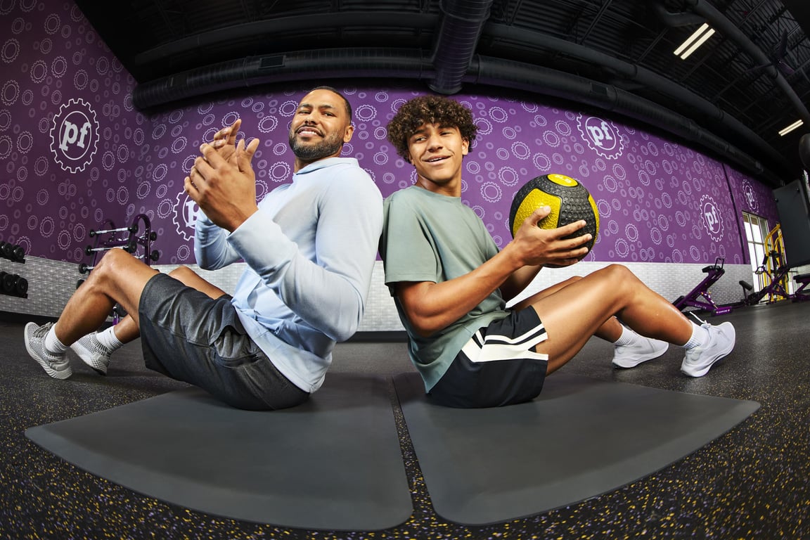 Teen and adult exercising together with a medicine ball at Planet Fitness, sitting back-to-back on mats in front of a purple gym wall.