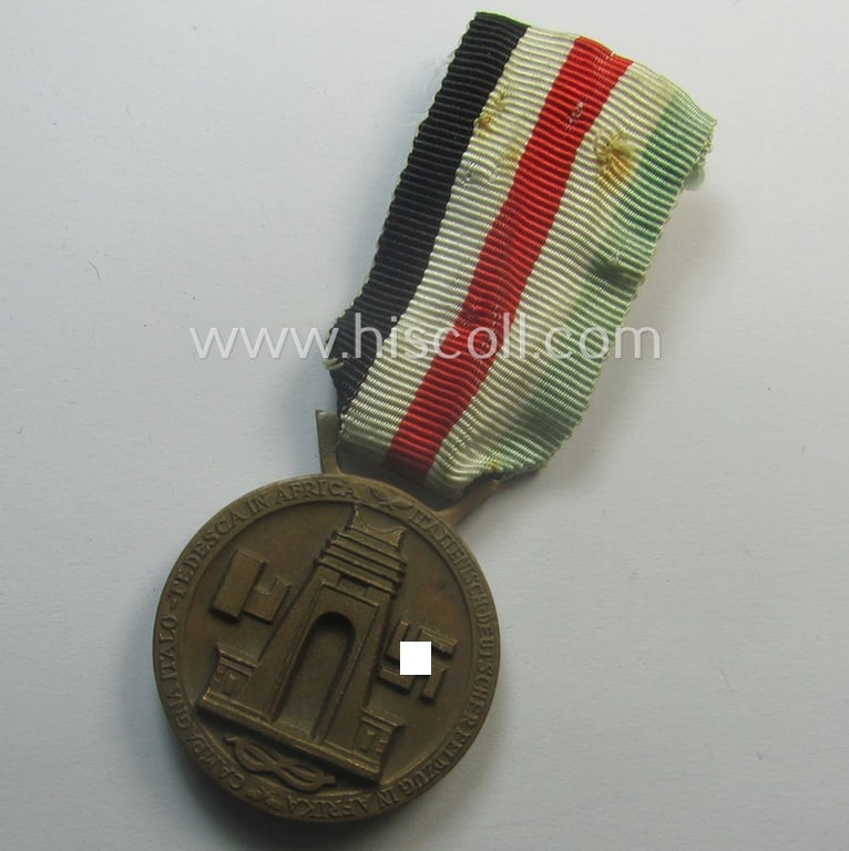Golden-bronze-coloured- (and I deem 'Buntmetall'-based-) example of a: 'Deutsch-Italienische Feldzugsmedaille' (or: German-Italian campaign-medal) that comes mounted onto its (regular-sized) piece of original (and somewhat faded!) ribbon